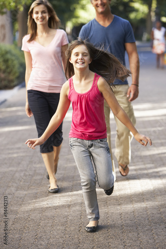 Fototapeta Naklejka Na Ścianę i Meble -  Daughter Runs Ahead Of Parents On Walk Through Summer Park