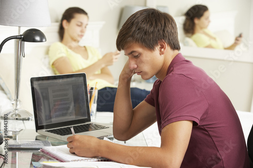 Teenage Boy Studying At Desk In Bedroom With Girl In Background © Monkey Business