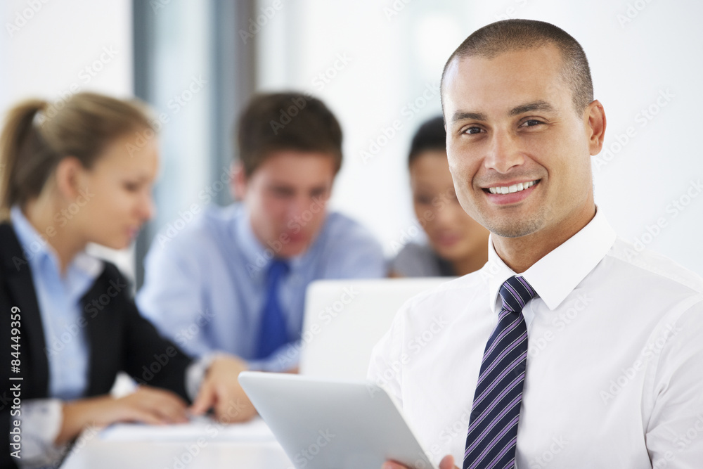 Portrait Of Male Executive Using Tablet Computer With Office Meeting In Background Portrait Of Male Executive Using Tablet Computer With Office Meeting In Background
