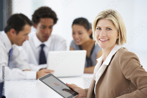 Portrait Of Female Executive Using Tablet Computer With Office Meeting In Background