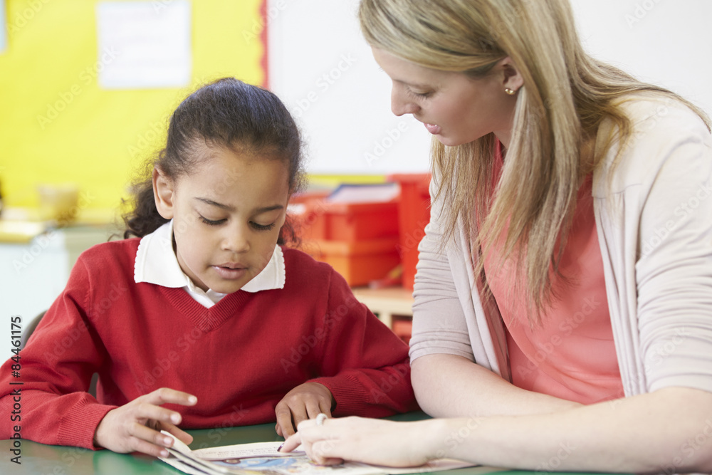 Teacher Helping Female Pupil With Practising Reading At Desk Teacher Helping Female Pupil With Practising Reading At Desk