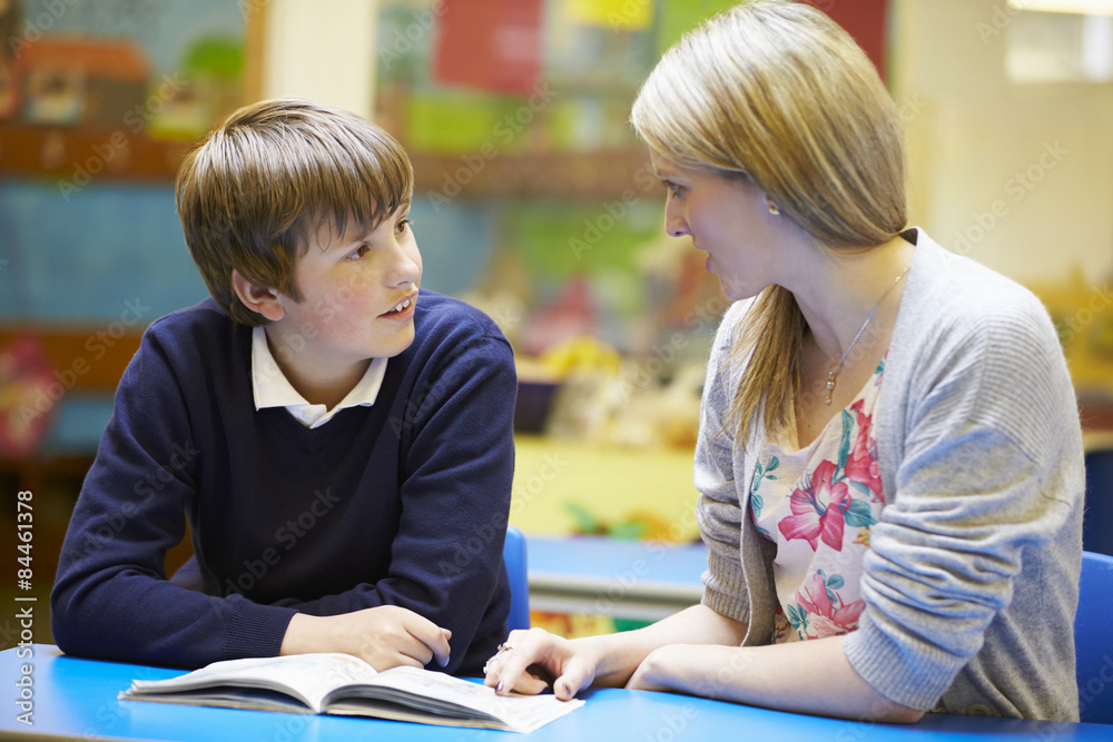 Teacher With Male Pupil Reading At Desk In Classroom Teacher With Male Pupil Reading At Desk In Classroom