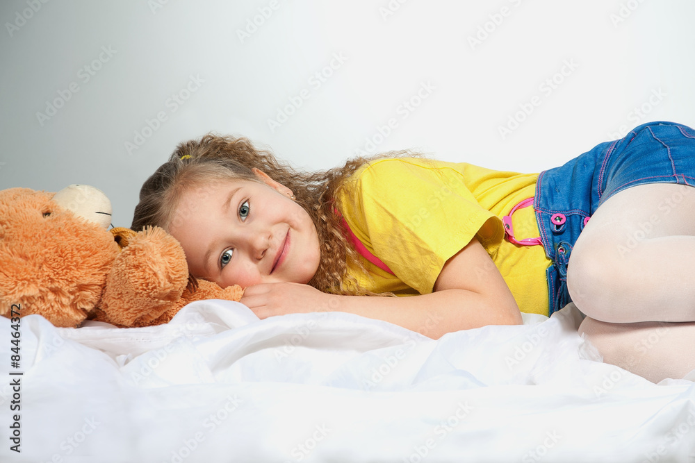 The smiling little girl with a teddy bear is lying on a white clot The smiling little girl with a teddy bear is lying on a white clot