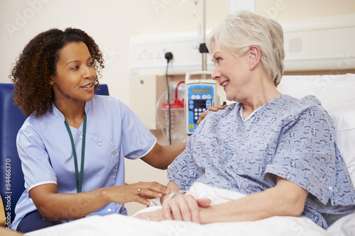 Nurse Sitting By Female Patient's Bed In Hospital