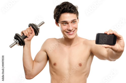 Skinny man taking a selfie with his phone while training his bicep muscle. Beautiful teenager lifting a dumbbell and taking a photo for facebook.Anorexic young man training to become stronger