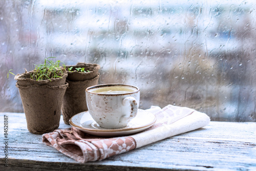 Fotografia Cup with warm drink on wooden table in front of window with rain drops, rainy weather