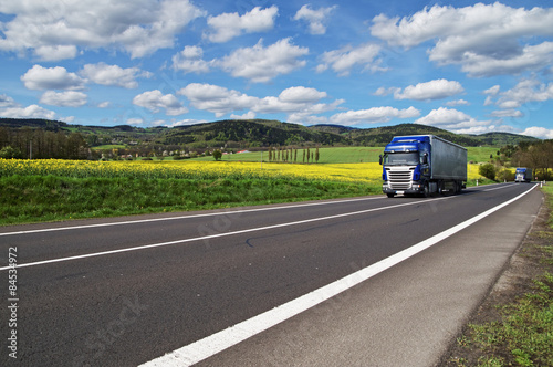 Obraz na płótnie Two blue trucks driving on asphalt road between yellow flowering rapeseed w salonie