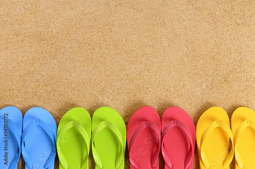 Row line of several pairs colorful flip flop sandals on a sand beach background lower border space for copy text photo
