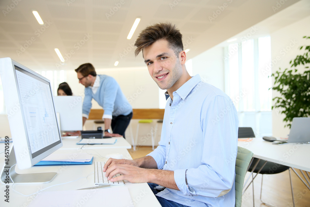 Office worker sitting in front of desktop, colleagues in background Office worker sitting in front of desktop, colleagues in background