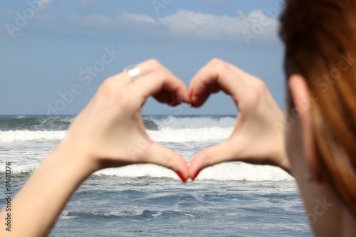 Female hand making a heart shape, ocean's waves in the background 
