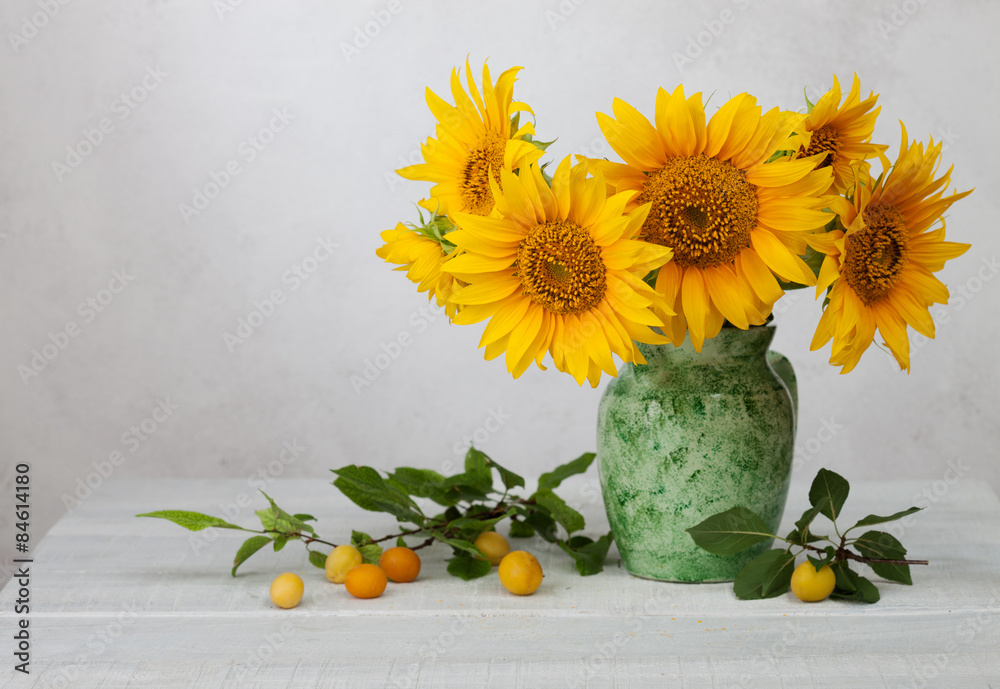 Obraz premium Bouquet of sunflowers in old ceramic jug against a white wooden wall. In the foreground branches with ripe cherry plum
