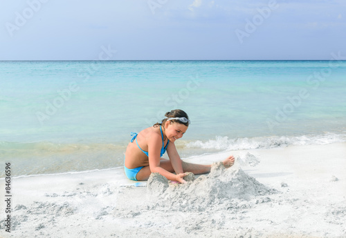 Little beautiful girl sitting and playing on white sand gorgeous beach against tranquil ocean and blue sky background