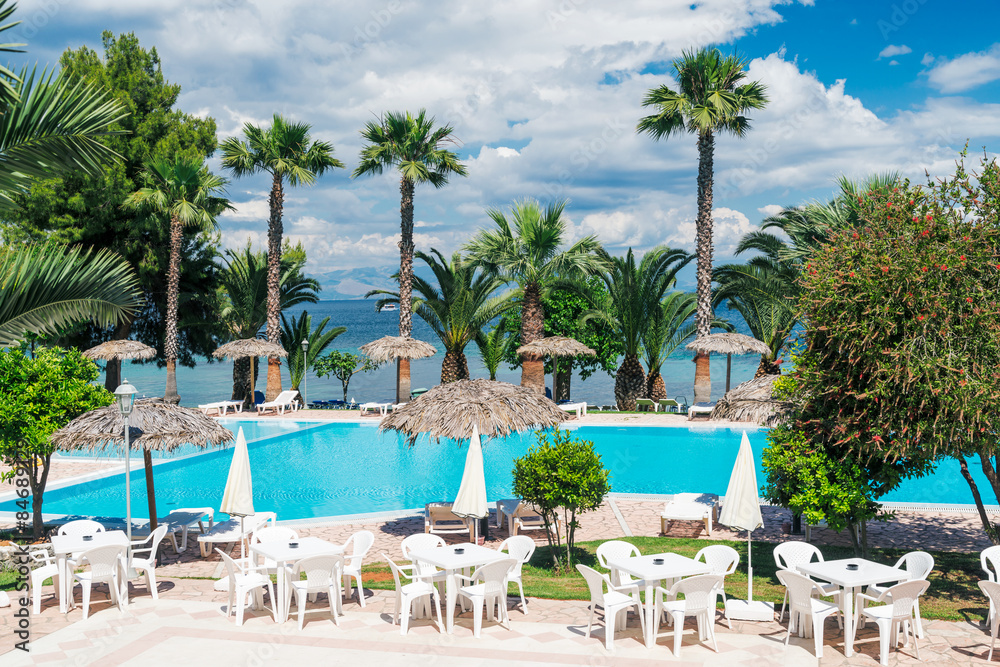 Palm trees, beach sunbeds and umbrellas near the pool by the sea in sunny day, Corfu, Greece.