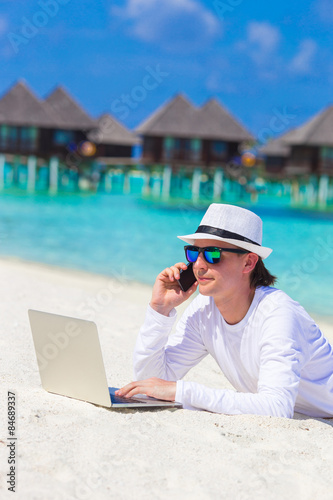 Young business man with computer and cell phone on white beach