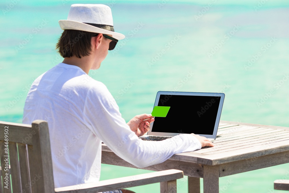 Young man working on laptop with credit card during tropical