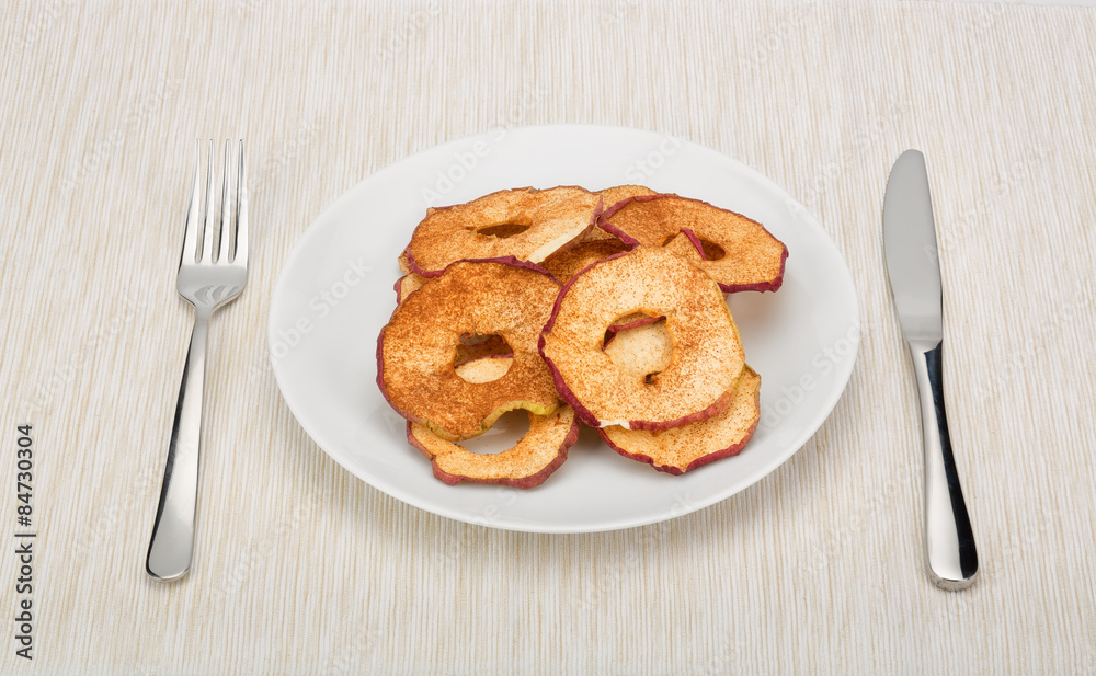 Dried apple slices sprinkled with cinnamon served on a white plate on tablecloth. Cutlery on both sides. 
