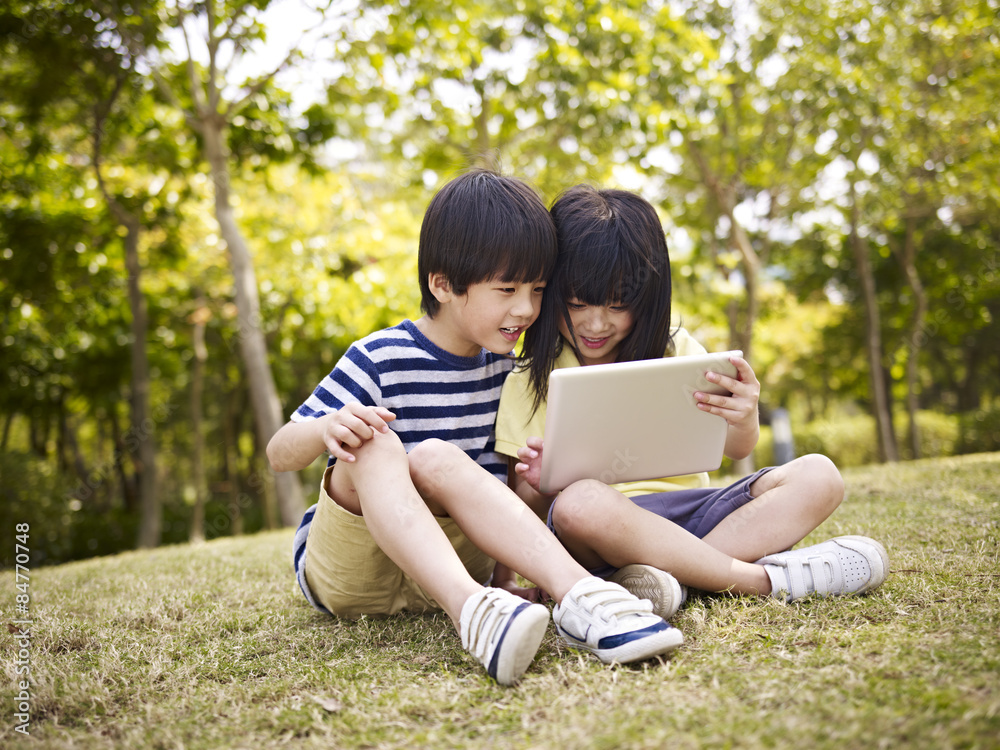 two asian children sitting on grass using tablet computer two asian children sitting on grass using tablet computer
