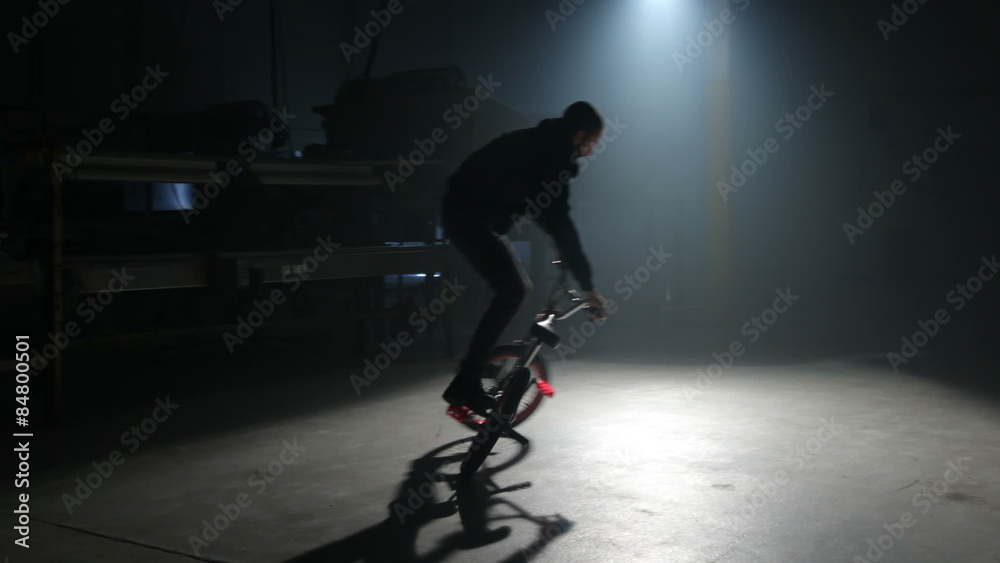 BMX rider doing tricks in dark warehouse.