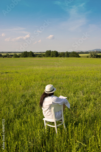 Woman from Behind Sitting on White Chair on Meadow Field and Drawing a Village