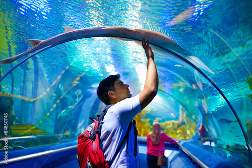 young man, tourist touching the glass under cramp-fish, while visiting marine underwater tunnel