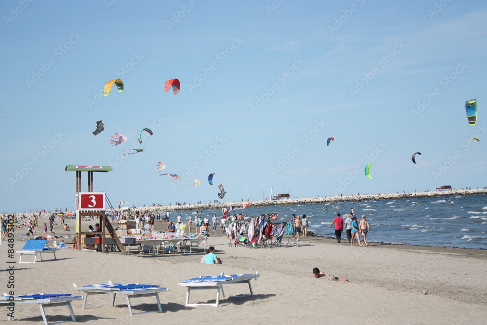 La foto rappresenta una spiaggia, nel tardo pomeriggio, con molti windsurfer tra le onde