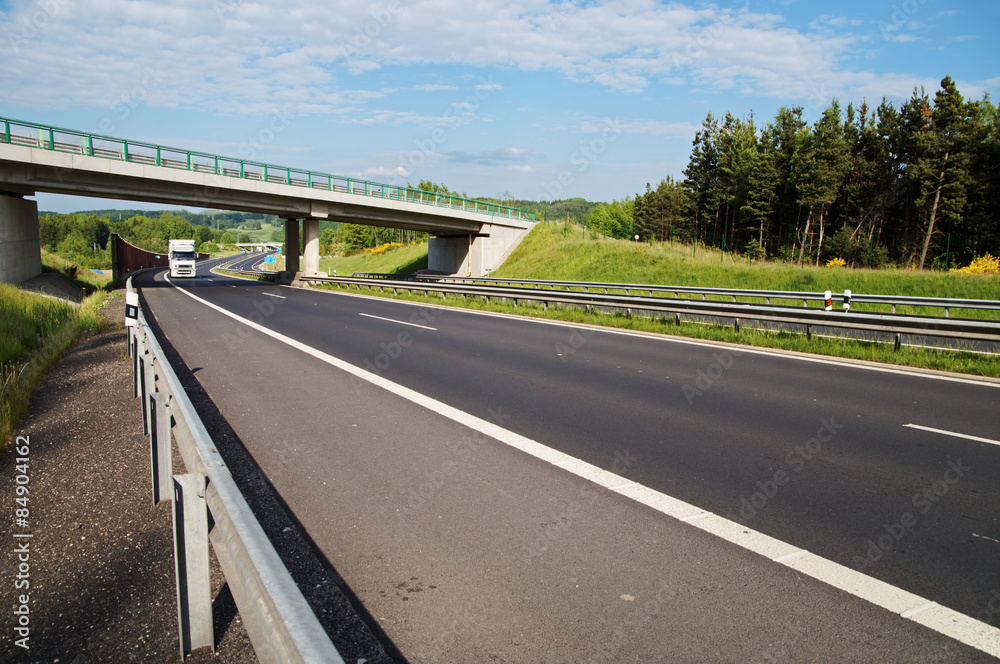 White truck entering under a concrete bridge over a highway in a wooded landscape