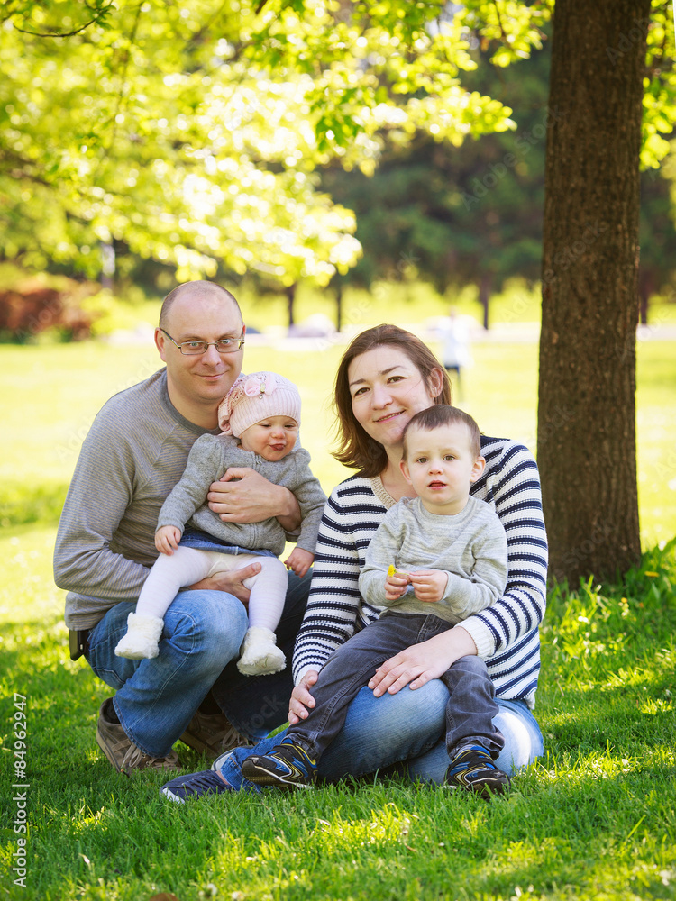 Fototapeta premium Portrait of mother, father, daughter and son, happy family of four, sitting on the meadow in the park outside on a spring summer bright day