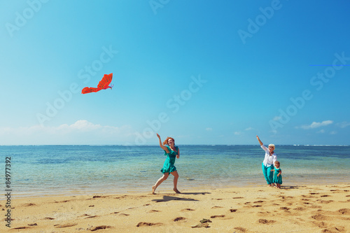 Carefree mother having a fun running with flying red kite on sea beach to baby girl and grandmother. Three generations of family, happy parenting and active leisure during summer vacation with child