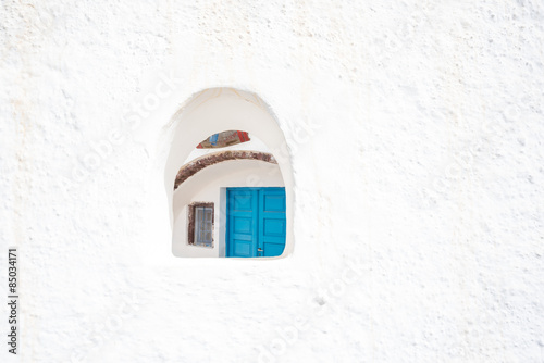 The blue door of a canava, or winery seen through a small window opening in a white wall in Oia, Santorini.