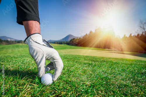 Hand with a glove is placing a golf ball on the ground.