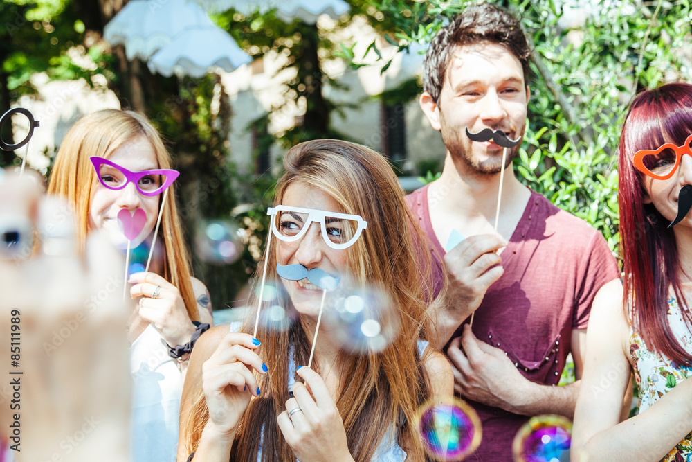Party! A group of friends, three women and a man have fun at a party in a park with a mustache and fake glasses, joking and talking to each other and playing with soap bubbles in the air! - obrazy, fototapety, plakaty 