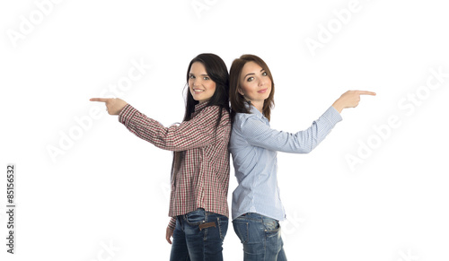 Two smiling girls pointing to opposite directions. Two young women stay back to back and pointing hands into opposite directions on white background casually dressed jeans style
