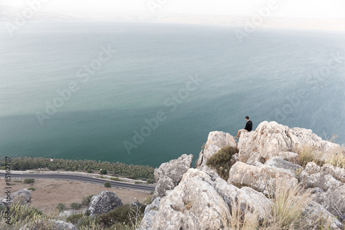 Fotografie Man sitting stone cliff above lake sea.