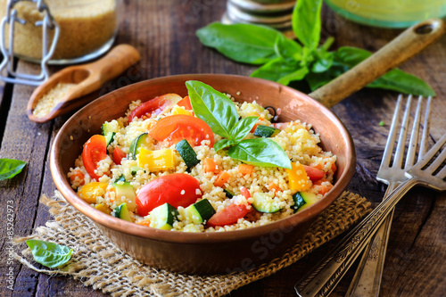 Delicious homemade vegetarian couscous with tomatoes, carrots, zucchini, yellow bell pepper and fresh basil on a dark rustic wooden kitchen table