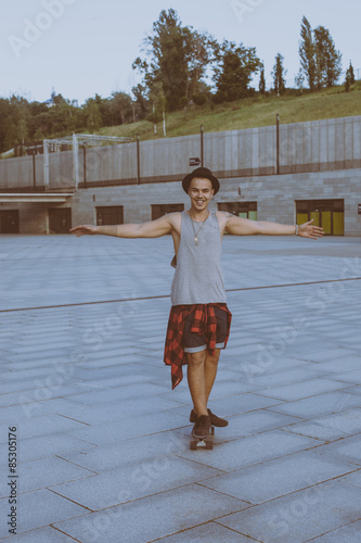 cool young and handsome caucasian brunette hipster skater guy wearing a hat posing smiling and having fun outside while skating with his skateboard summer day in the city