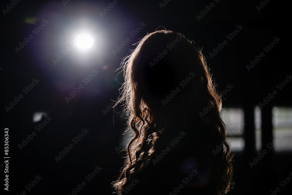 Dunkelheit / Eine Frau steht in einem dunklen Raum und wird von einem Scheinwerfer von hinten angeleuchtet. Zu sehen ist nur ihre Silhouette. - obrazy, fototapety, plakaty 