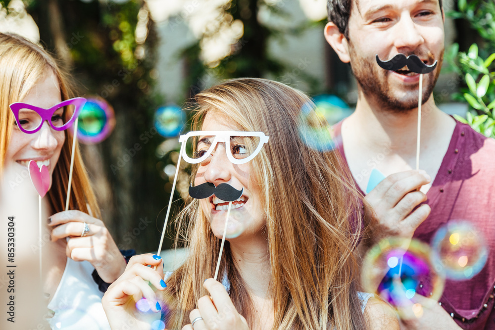 Party! A group of friends, two women and a man have fun at a party in a park with a mustache and fake glasses, joking and talking to each other and playing with soap bubbles in the air! - obrazy, fototapety, plakaty 