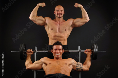 Two muscular man posing with dumbells on dark background