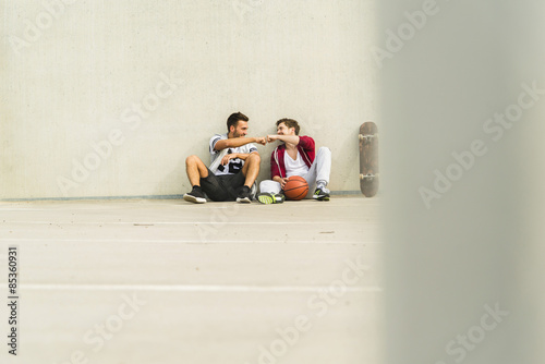 Two young men sitting with skateboard and basketball on parking level