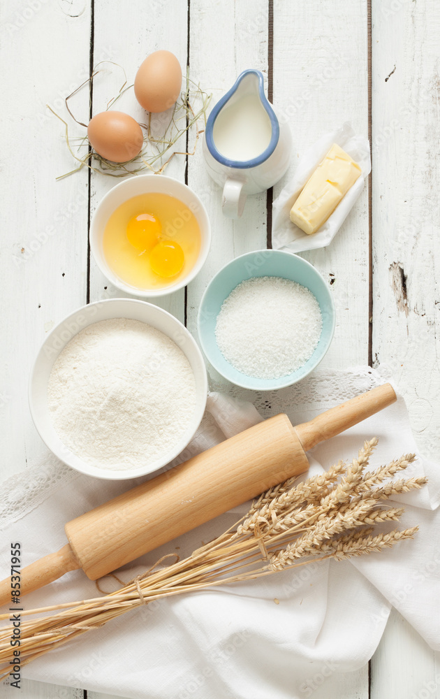 Baking cake in rustic kitchen - dough recipe ingredients (eggs, flour, milk, butter, sugar) on white planked wooden table from above.
