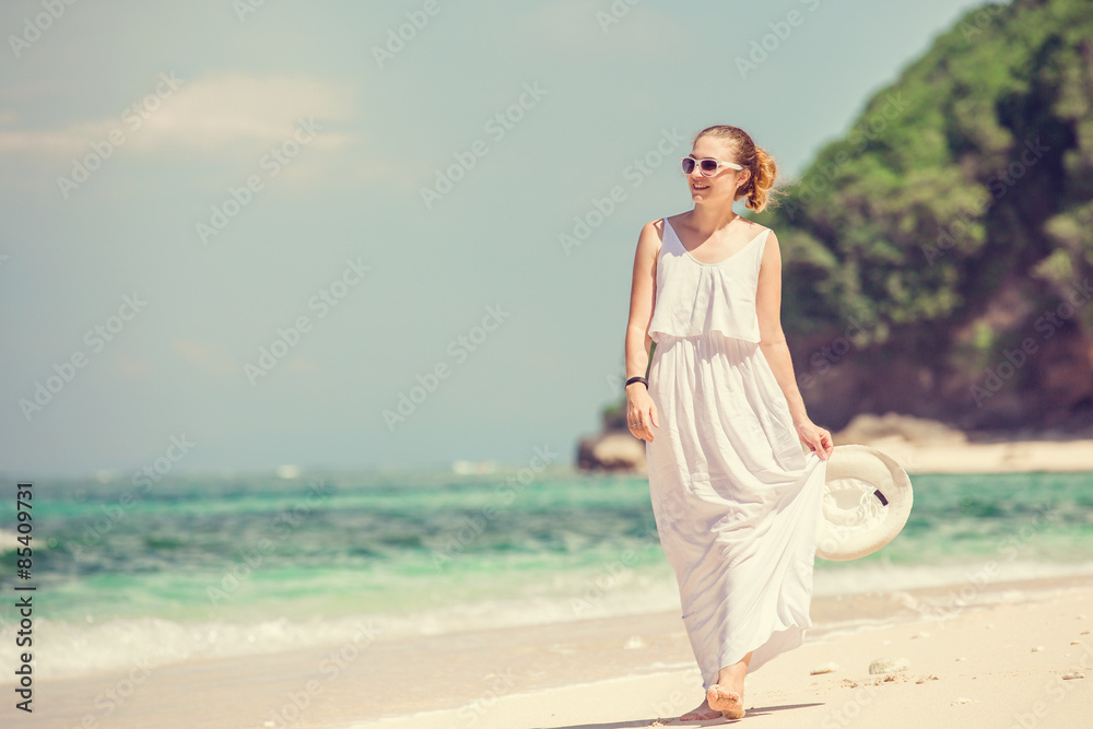 Young woman in long white dress holding hat walks along tropical beach having great summer time on holidays Young woman in long white dress holding hat walks along tropical beach having great summer time on holidays