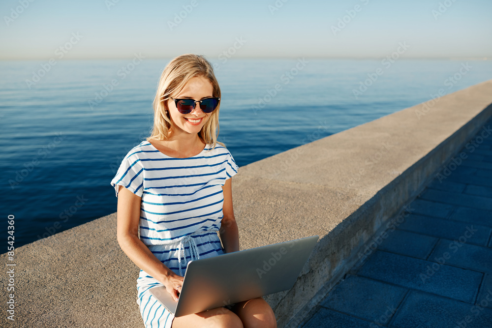 Beautiful young woman sitting on beach with laptop smiling