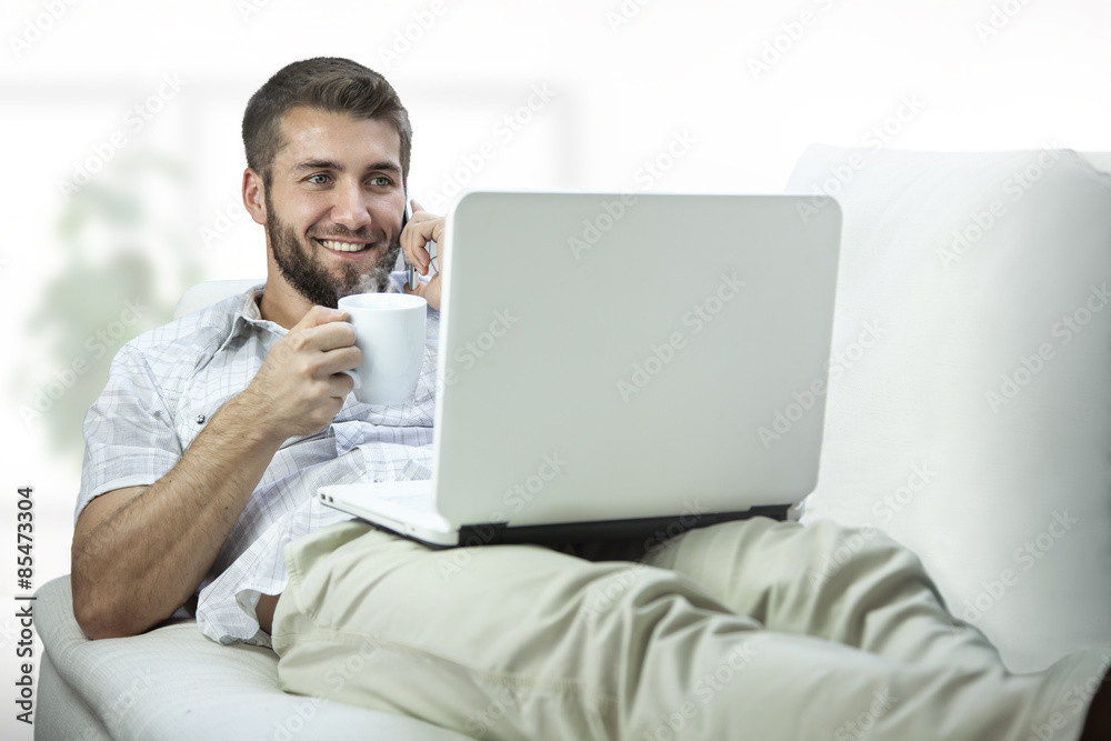 Attraktiver Mann liegt auf der Couch mit Notebook und telefoniert