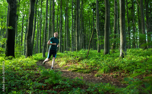 young man easy running on the trail in the forest with green leaves