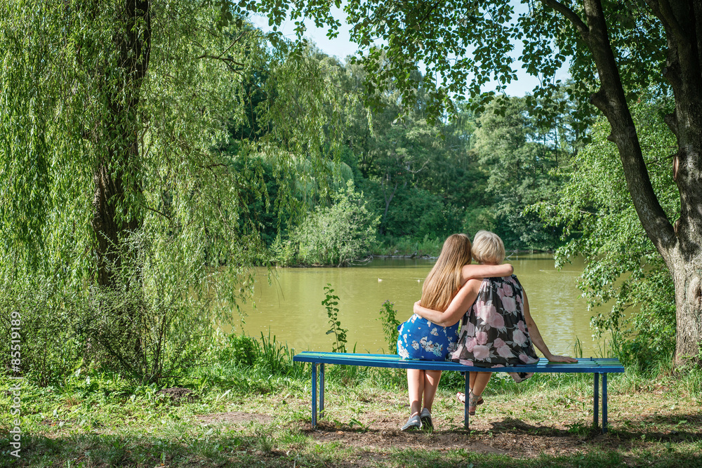 Two women of different generations sitting on a bench near a pond in the summer. Mother and daughter hugging. Grandmother and granddaughter. Two women of different generations sitting on a bench near a pond in the summer. Mother and daughter hugging. Grandmother and granddaughter.