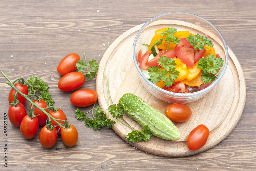 Salad of fresh yellow and pink tomatoes and cucumber with parsley in a glass bowl and vegetables on a wooden table Salad of fresh yellow and pink tomatoes and cucumber with parsley in a glass bowl and vegetables on a wooden table