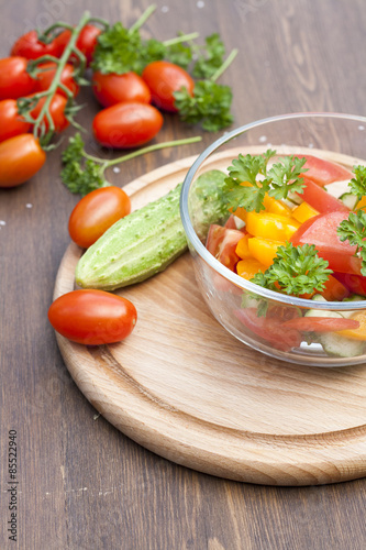 Salad of fresh yellow and pink tomatoes and cucumber with parsley in a glass bowl and vegetables on a wooden table