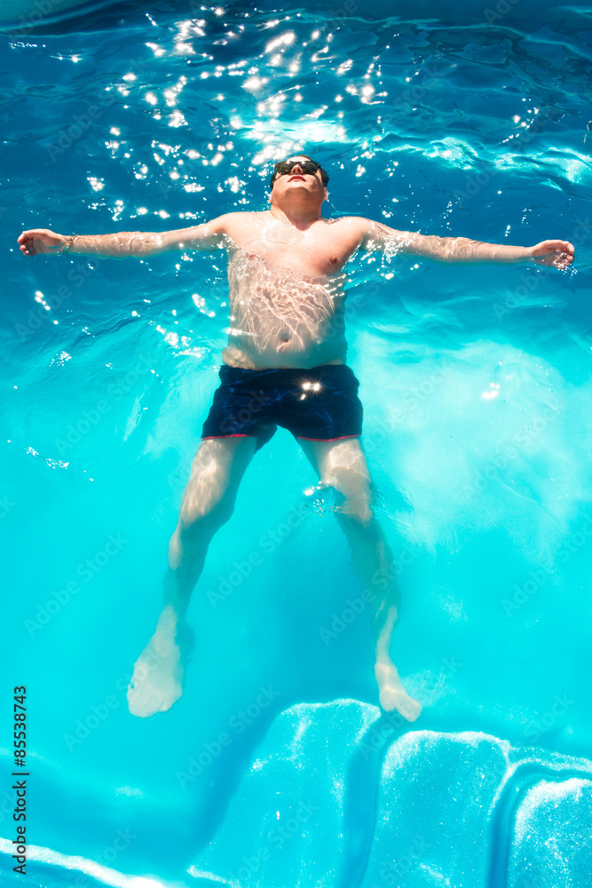 Man in sunglasses is laying on blue water surface in swimming pool.