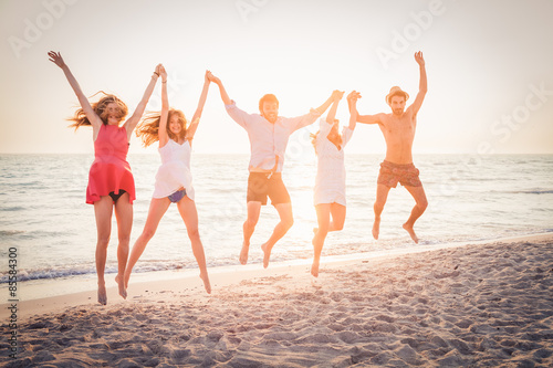 Summer beach at sunset. A group of five friends jumping in the air with his hands to the sky on the beach. Three women and two men jumping with sun at sunset behind them