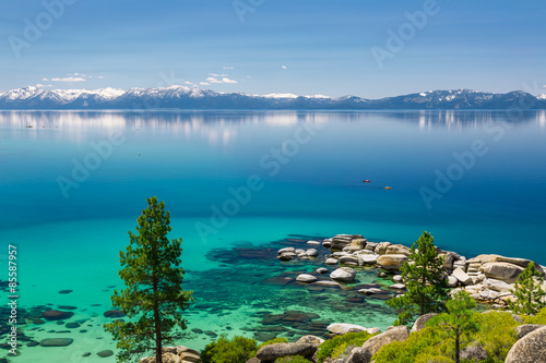 Lake Tahoe calm turquoise waters with view on Sierra Nevada snowy peaks. There are two kayaks close to shore rocks.